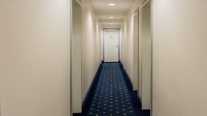 Un couloir étroit et bien éclairé, doté d’une moquette bleue et de murs blancs, qui mène à une porte portant le numéro 55.