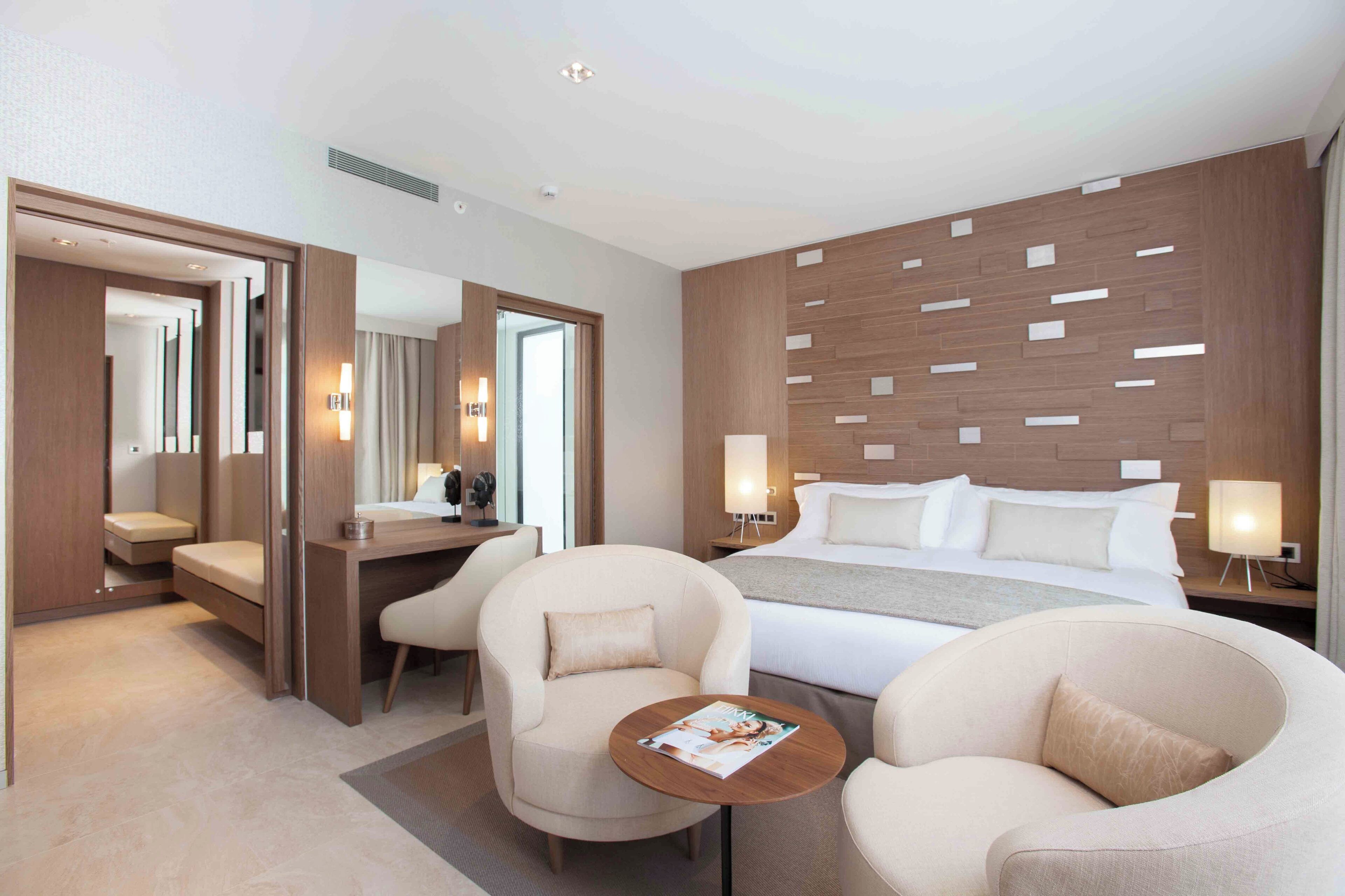 A modern hotel room with a bed, two armchairs, a desk, and a small table.