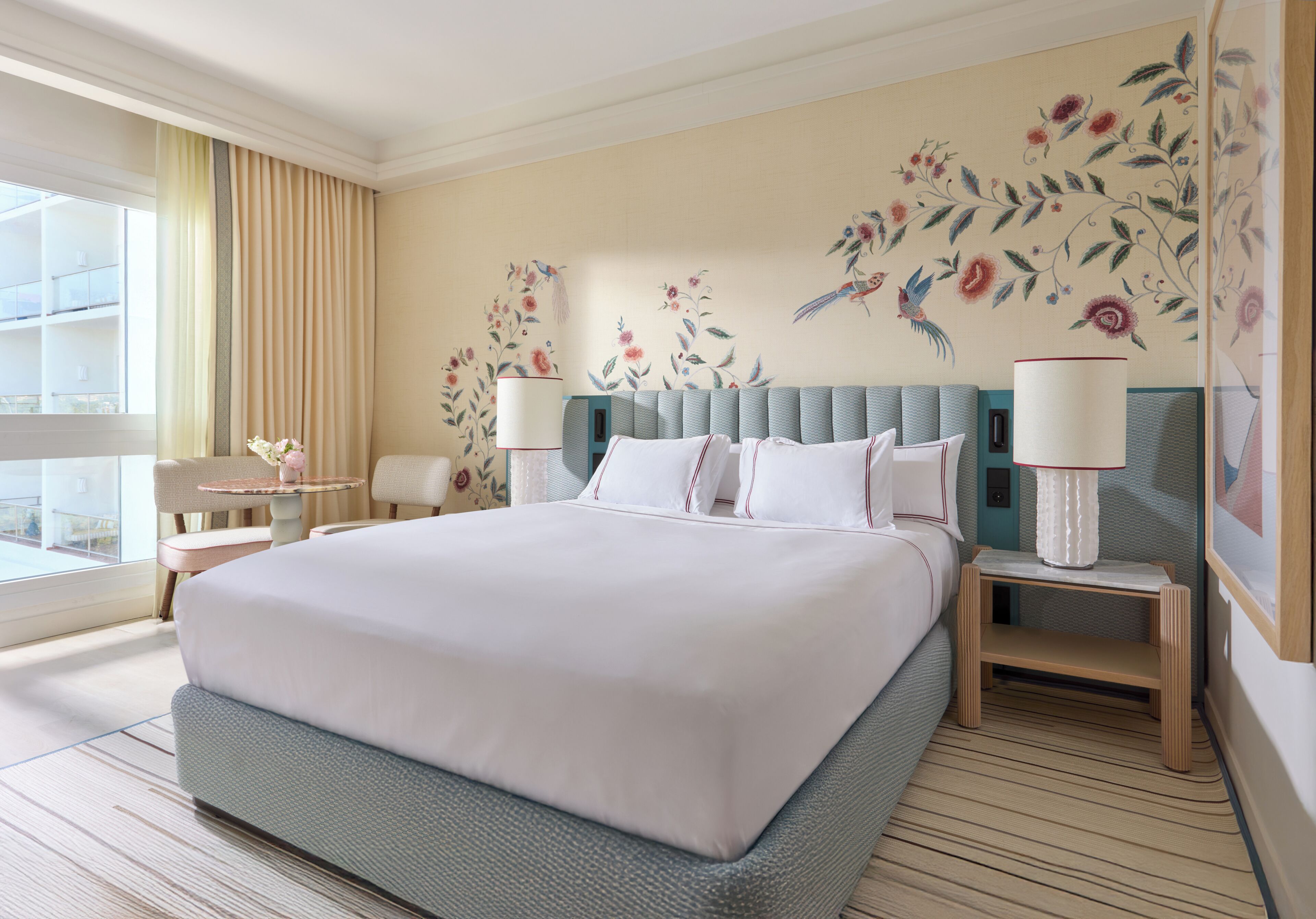 A hotel room with a large bed, two bedside tables, a window with curtains, and floral wall art.