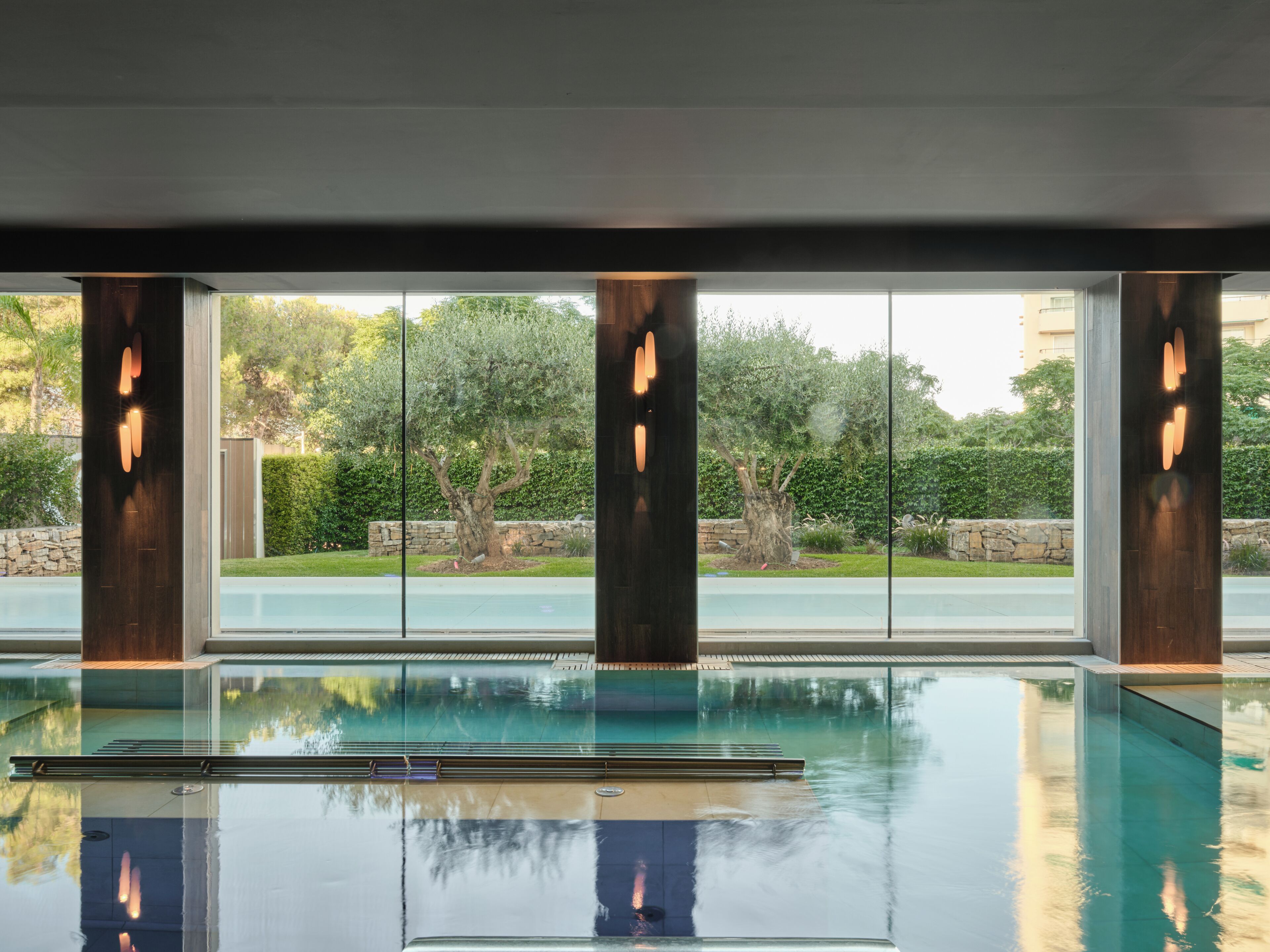 A modern indoor swimming pool with clear glass walls and wooden pillars, surrounded by a landscaped garden.