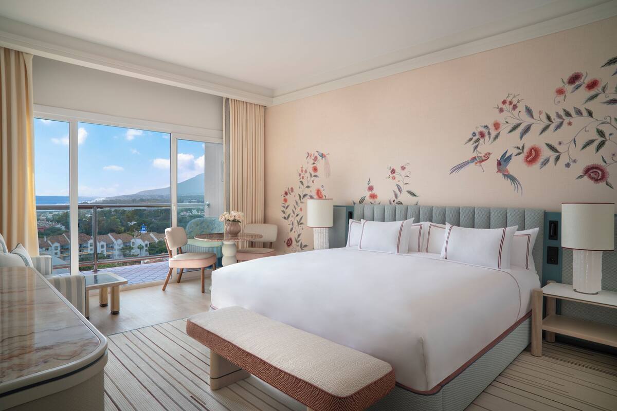 A hotel room with a large bed, a sitting area with a chair and table, a view of the outside, and a decorative wall.