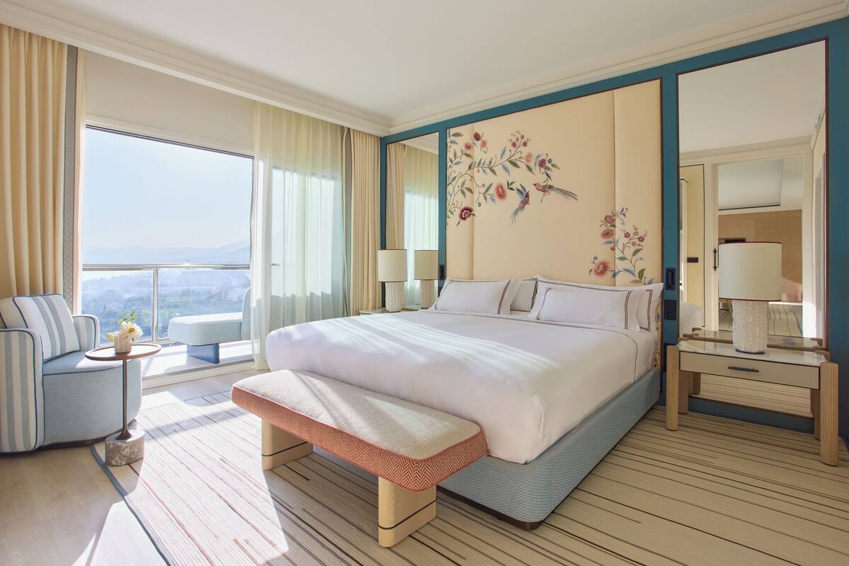 A hotel room with a large bed, a bench, a small table with a vase, a nightstand with a lamp, and a view of the outdoors.