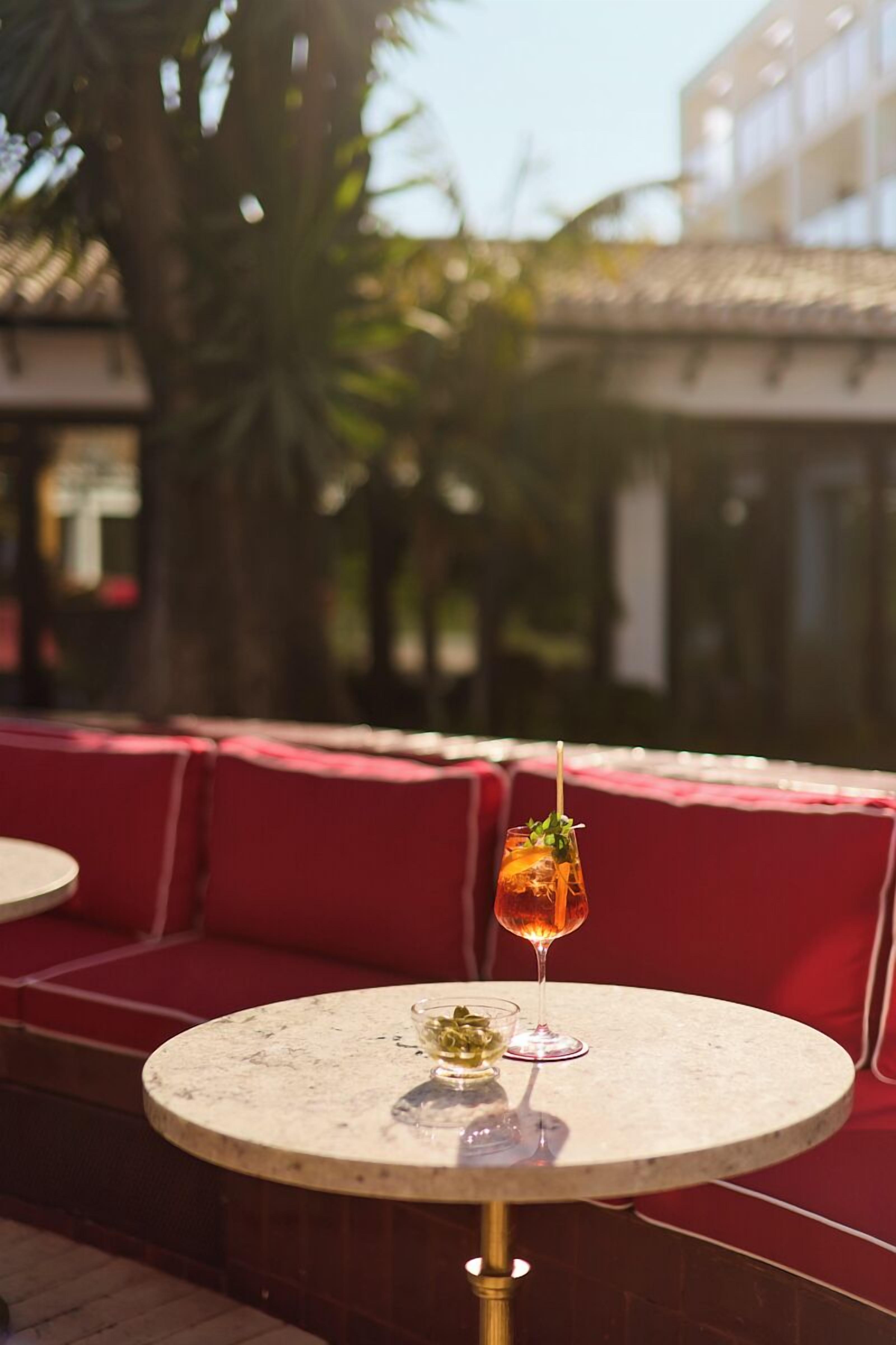 A round table with a glass of amber-colored drink and a small bowl of green garnish, set against a red cushioned bench in an outdoor seating area.