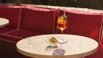 A round table with a glass of amber-colored drink and a small bowl of green garnish, set against a red cushioned bench in an outdoor seating area.