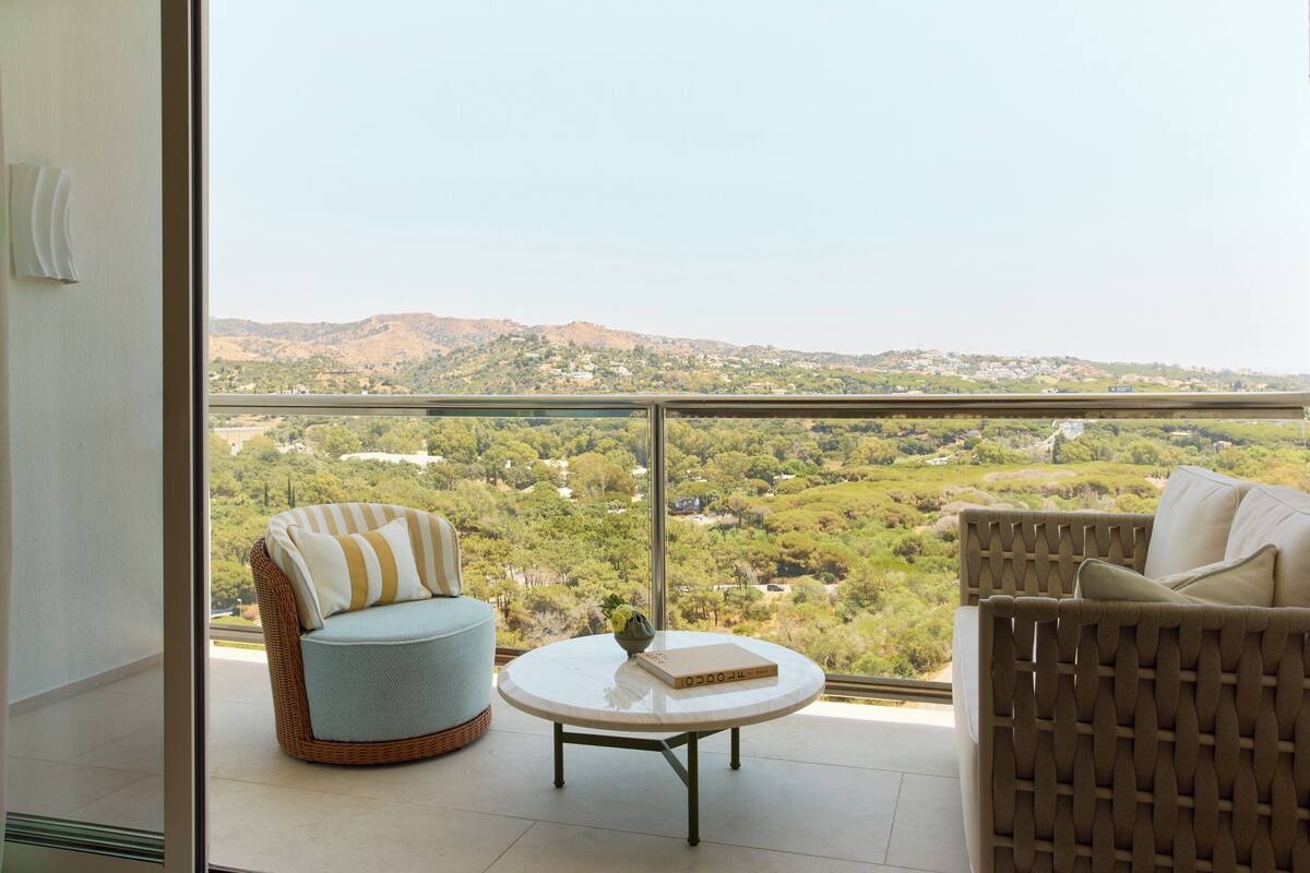 A balcony with wicker furniture, a round table, and a view of a hilly landscape.