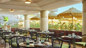 Een restaurant met terras, met rieten stoelen en tafels, en grote parasols voor schaduw.