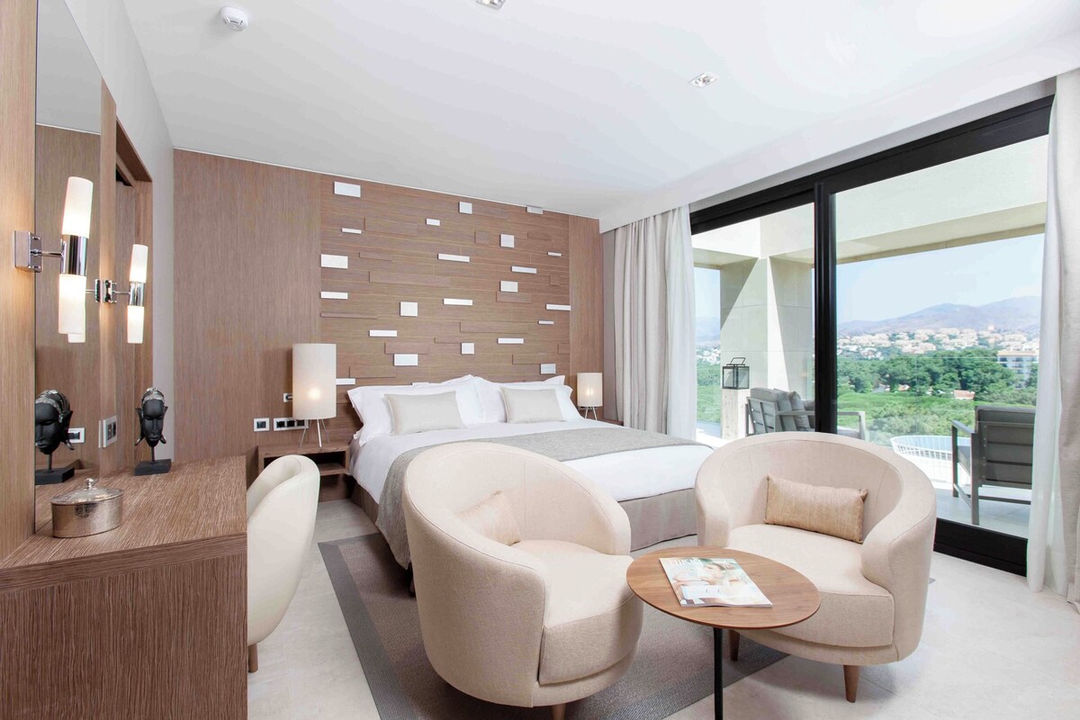A modern hotel room with a large bed, two armchairs, a small table, and a view of the city through large windows.