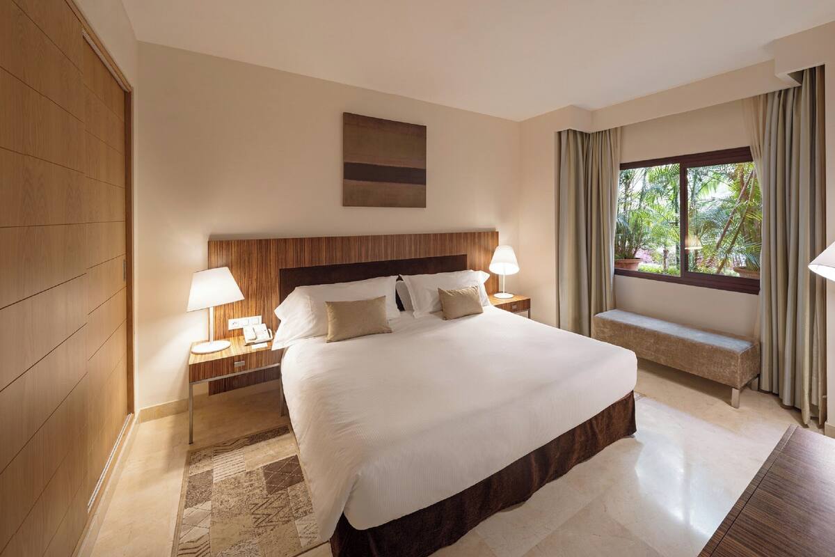 A hotel room with a large bed, bedside lamps, a bench, a window with curtains, and a view of greenery outside.