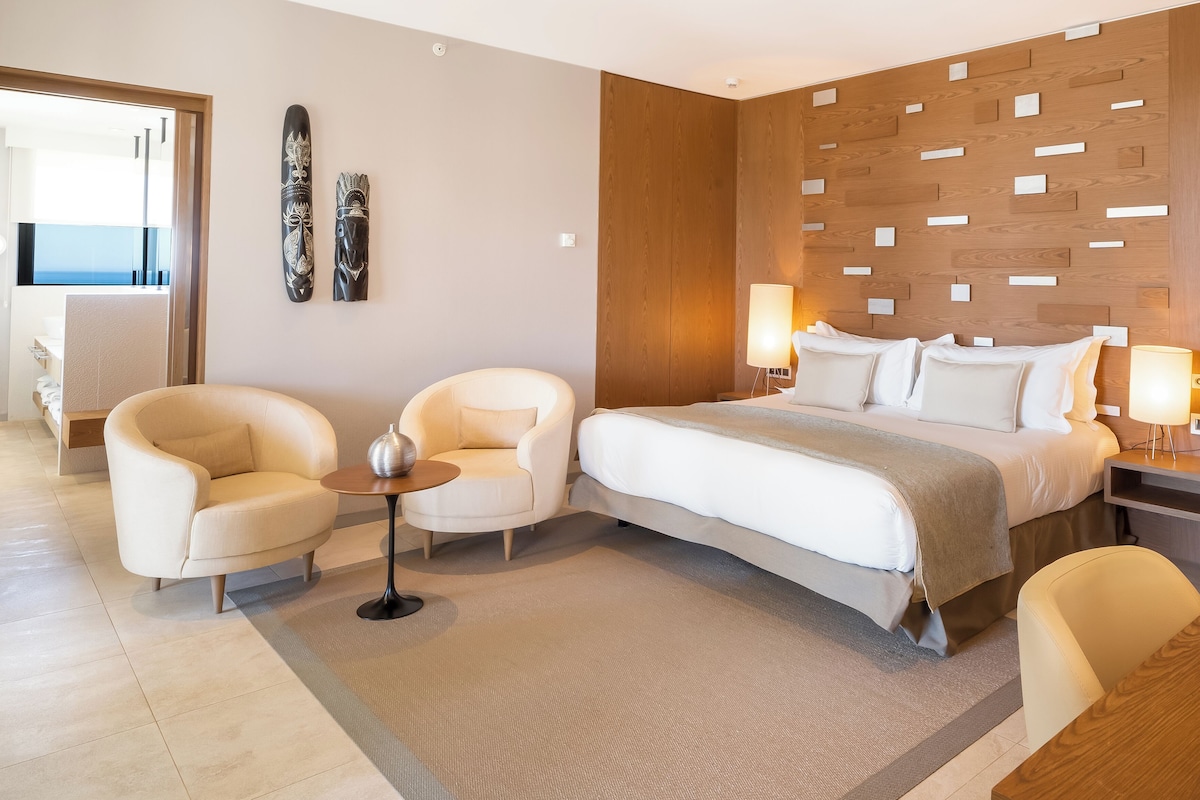A modern hotel room with a large bed, two armchairs, a small table, and a wooden accent wall.