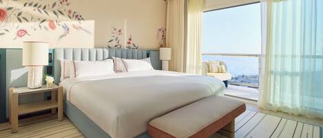 A hotel room with a large bed, a bench, a nightstand, a lamp, a wall mural, and a view of the city.