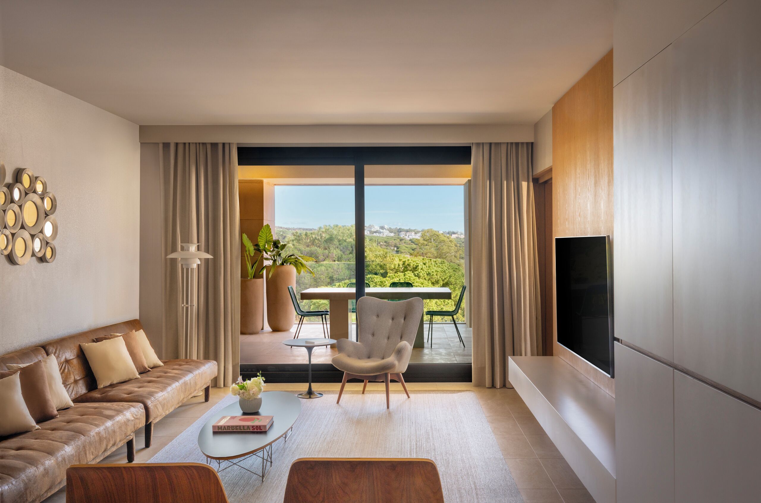 Un salon moderne avec un canapé, un fauteuil, une table basse et un téléviseur. Il y a un balcon avec une table et des chaises, ainsi qu’une grande fenêtre offrant une vue sur la ville.