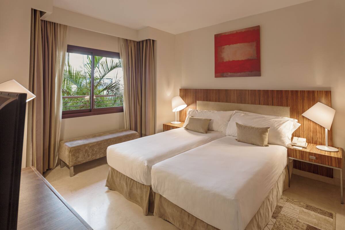 A hotel room with two beds, a bench, a desk, a TV, and a window with curtains.