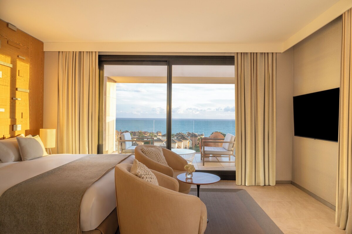 A hotel room with a bed, two armchairs, a small table, a TV, and a balcony with a view of the sea.