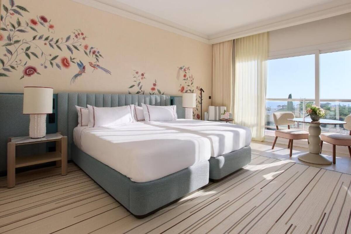 A hotel room with a large bed, two bedside tables, a chair, a small table with a vase, and a balcony with a view.