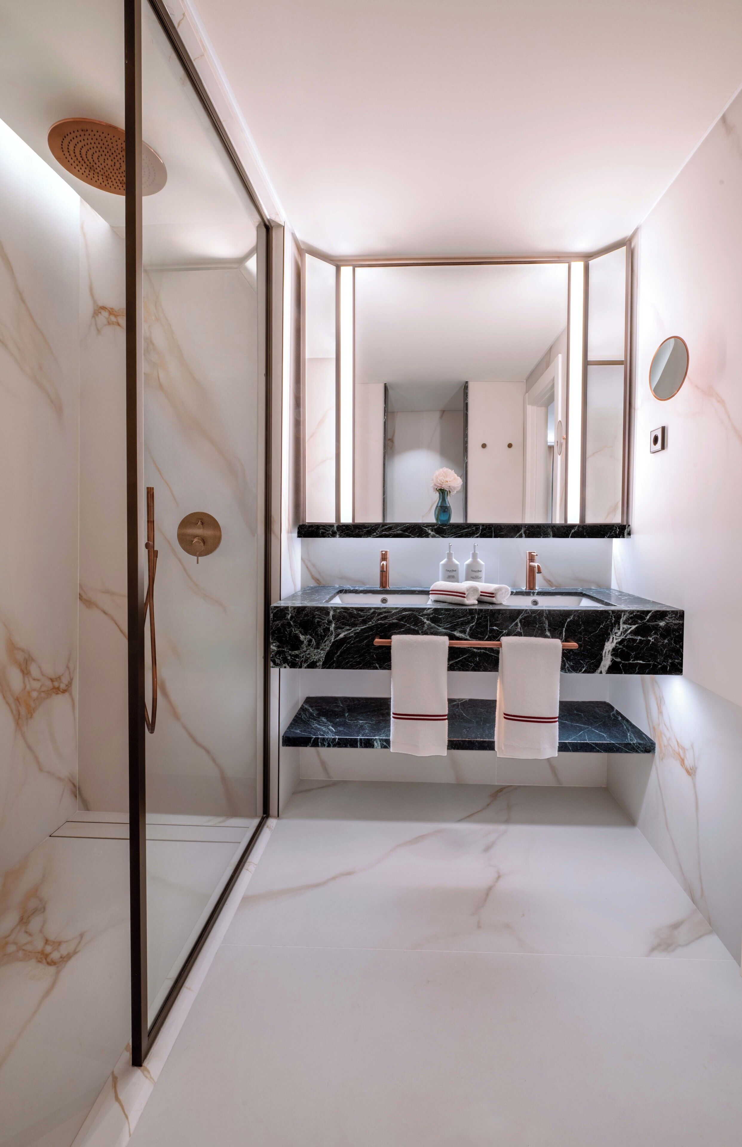 Een moderne badkamer met een douchecabine van glas, een marmeren vloer, een zwart marmeren aanrecht en een grote spiegel.