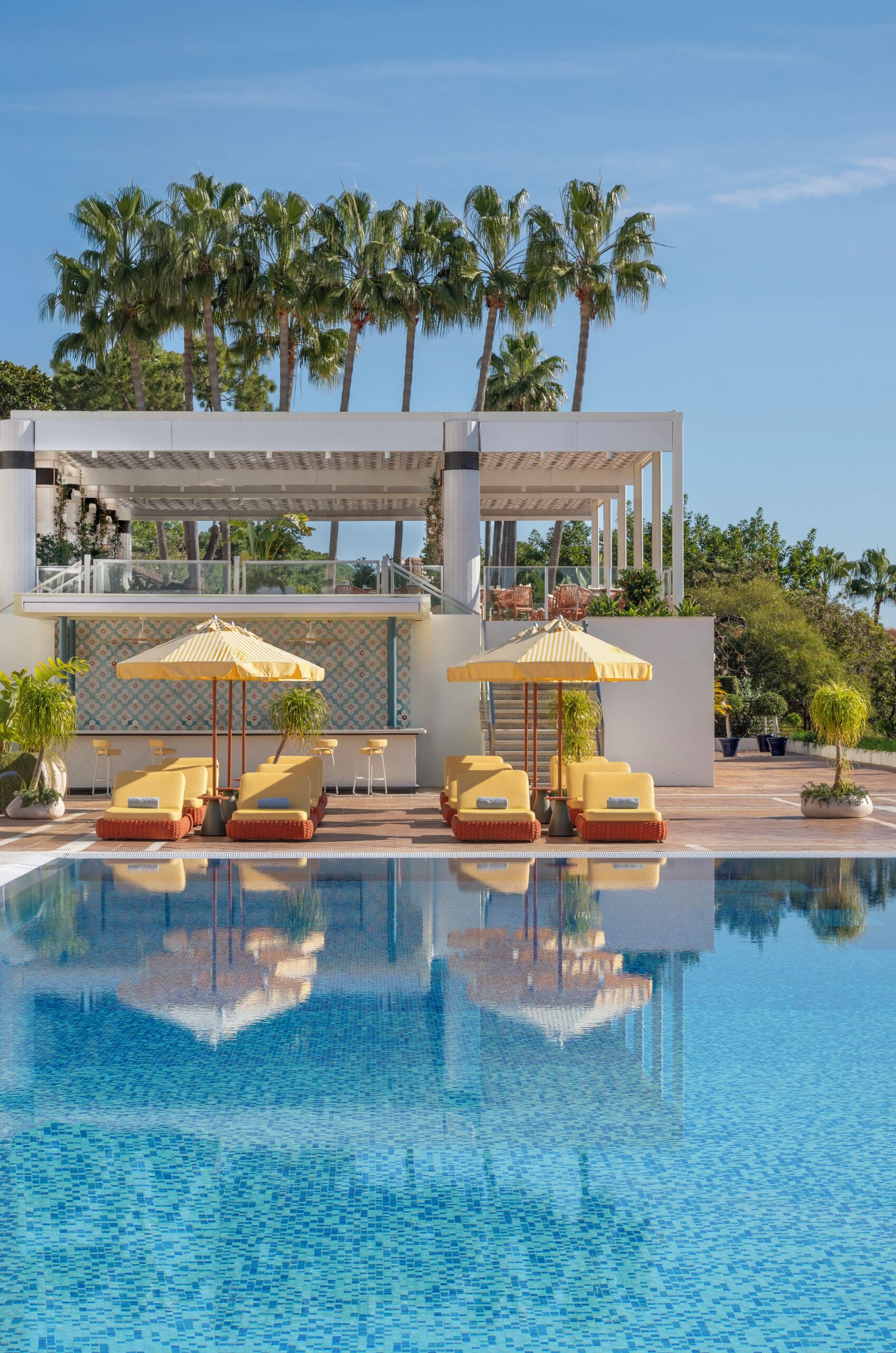 A pool area with lounge chairs and umbrellas, surrounded by palm trees and a modern building with a balcony.