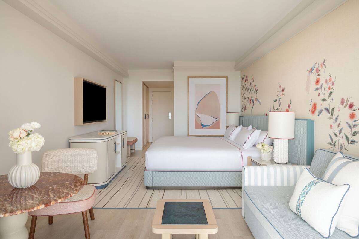 A hotel room with a bed, a sofa, a dining table, a TV, and a desk.