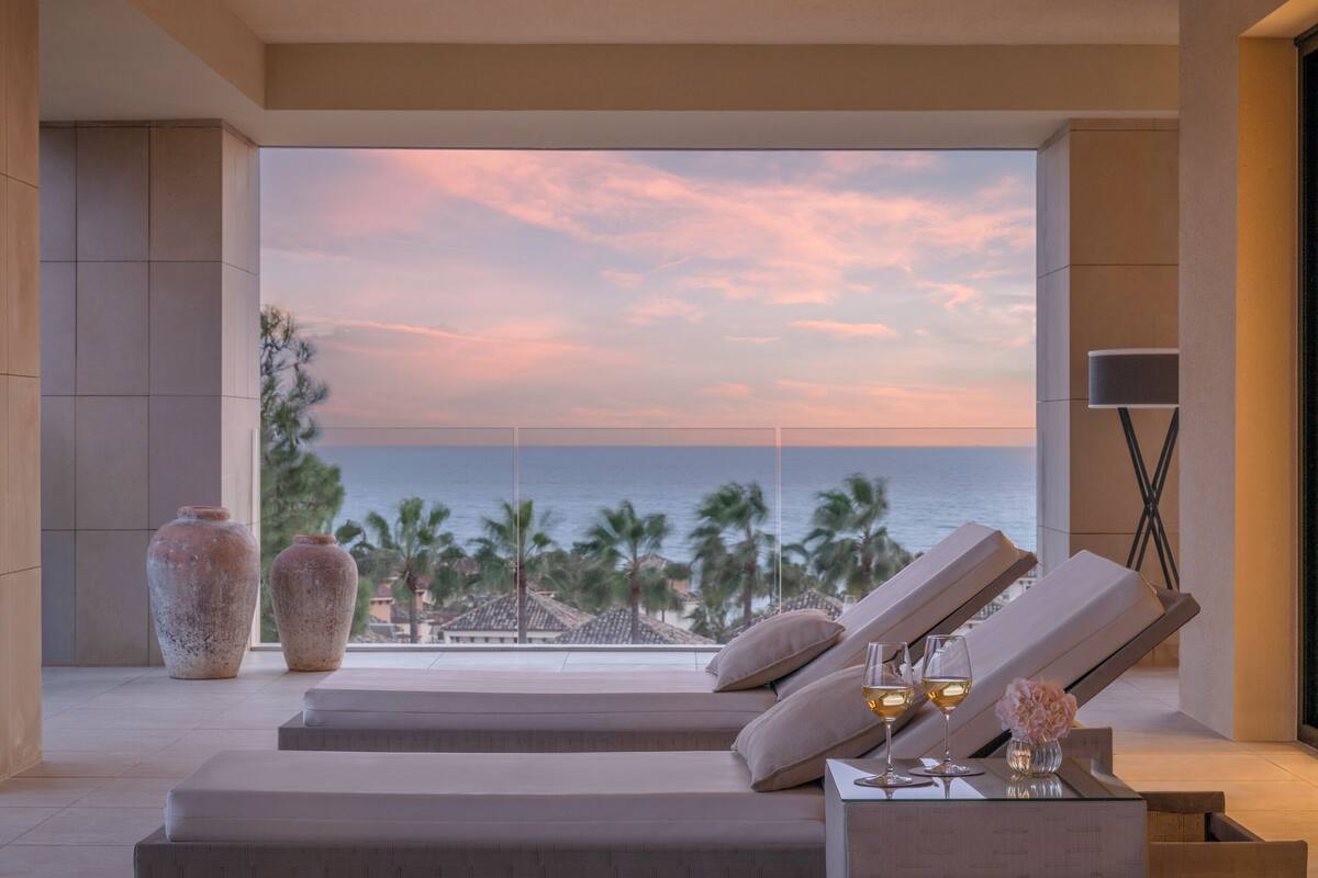 A modern lounge area with a view of the ocean at sunset, featuring reclining chairs, a small table with drinks, and decorative vases.