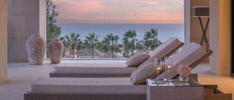 A modern lounge area with a view of the ocean at sunset, featuring reclining chairs, a small table with drinks, and decorative vases.