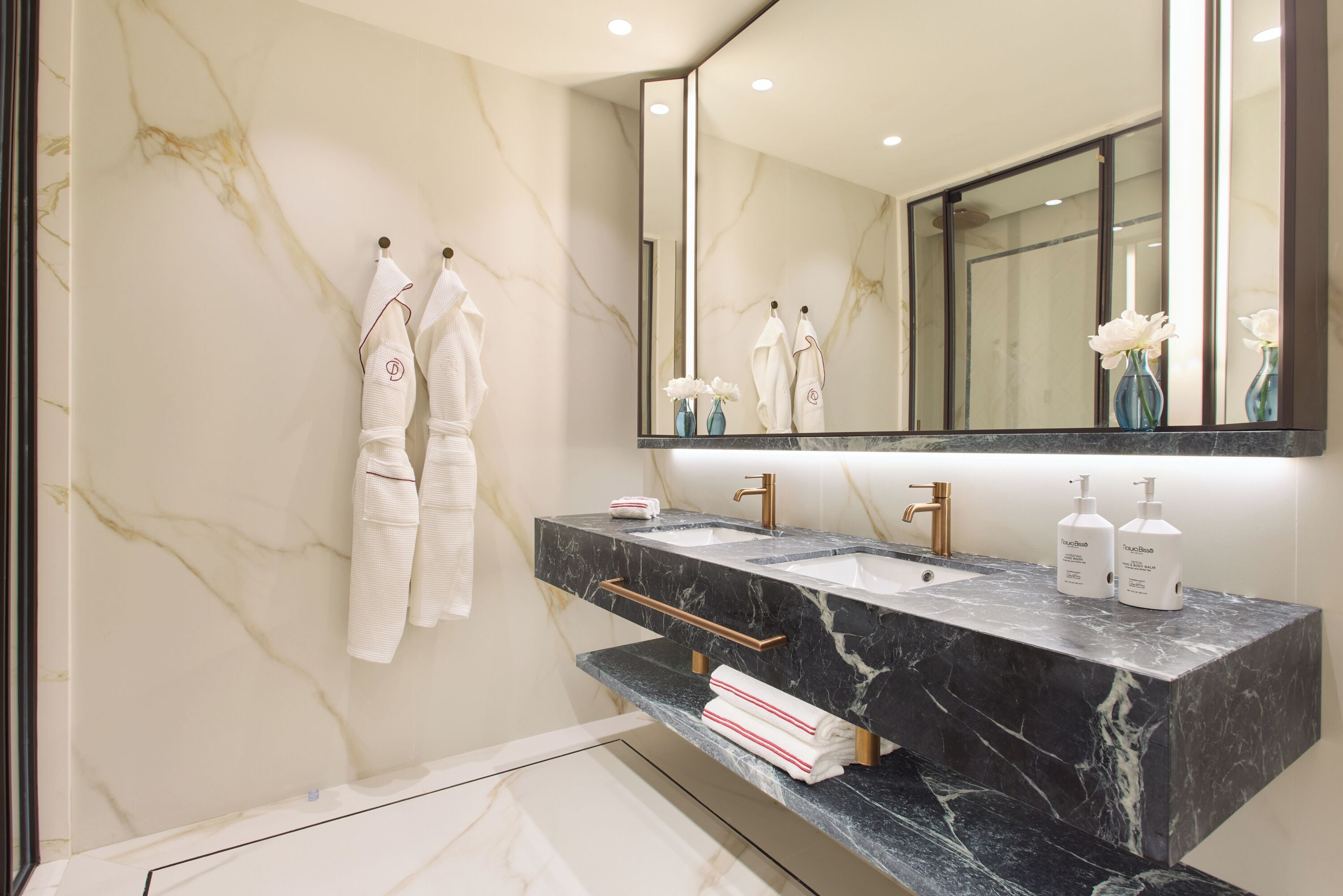 A modern bathroom with a marble wall, a large mirror, two sinks, and a towel rack.
