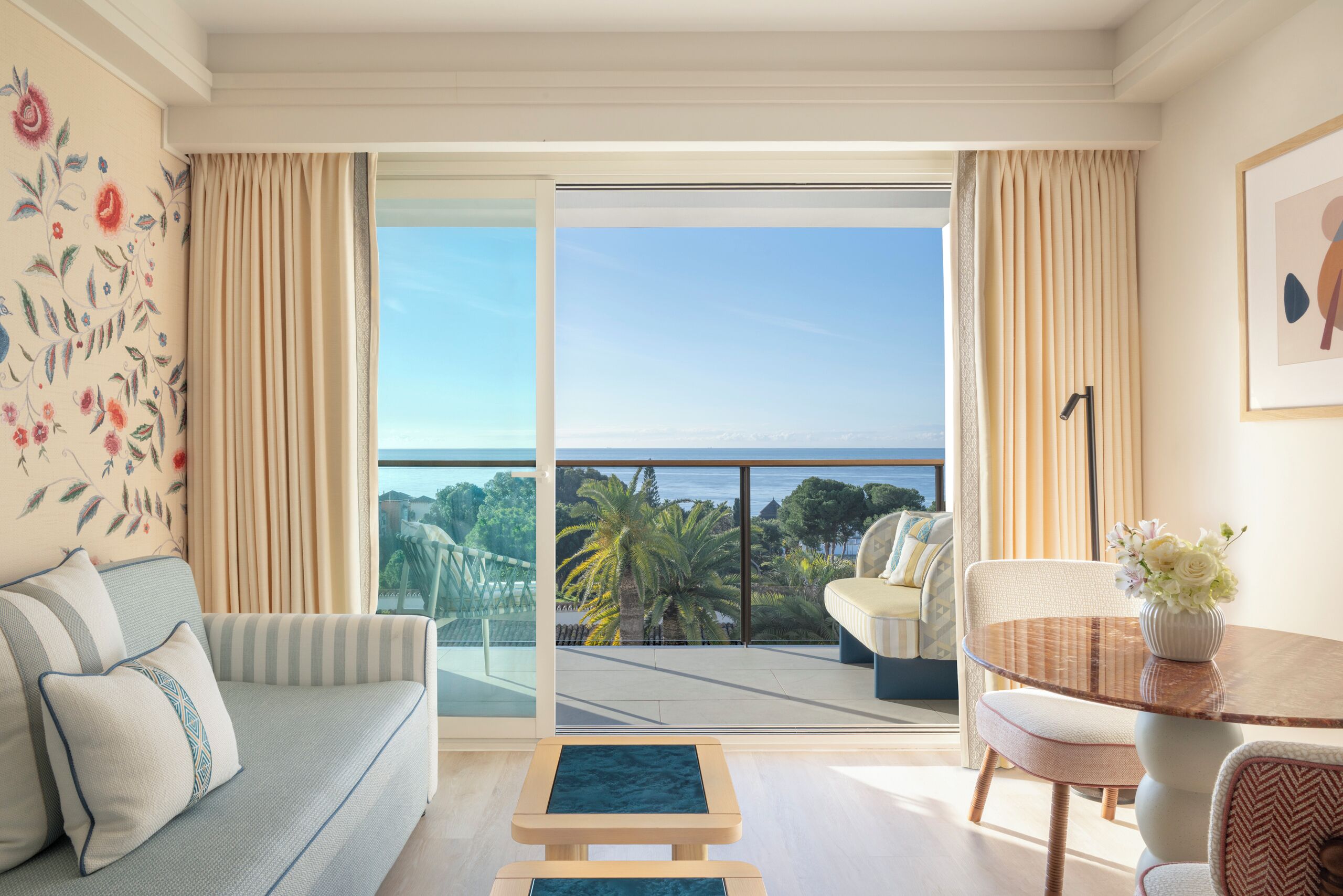 Una sala de estar moderna con un sofá, una mesita y una silla, un balcón con vista a palmeras y al mar, y un cuadro en la pared.