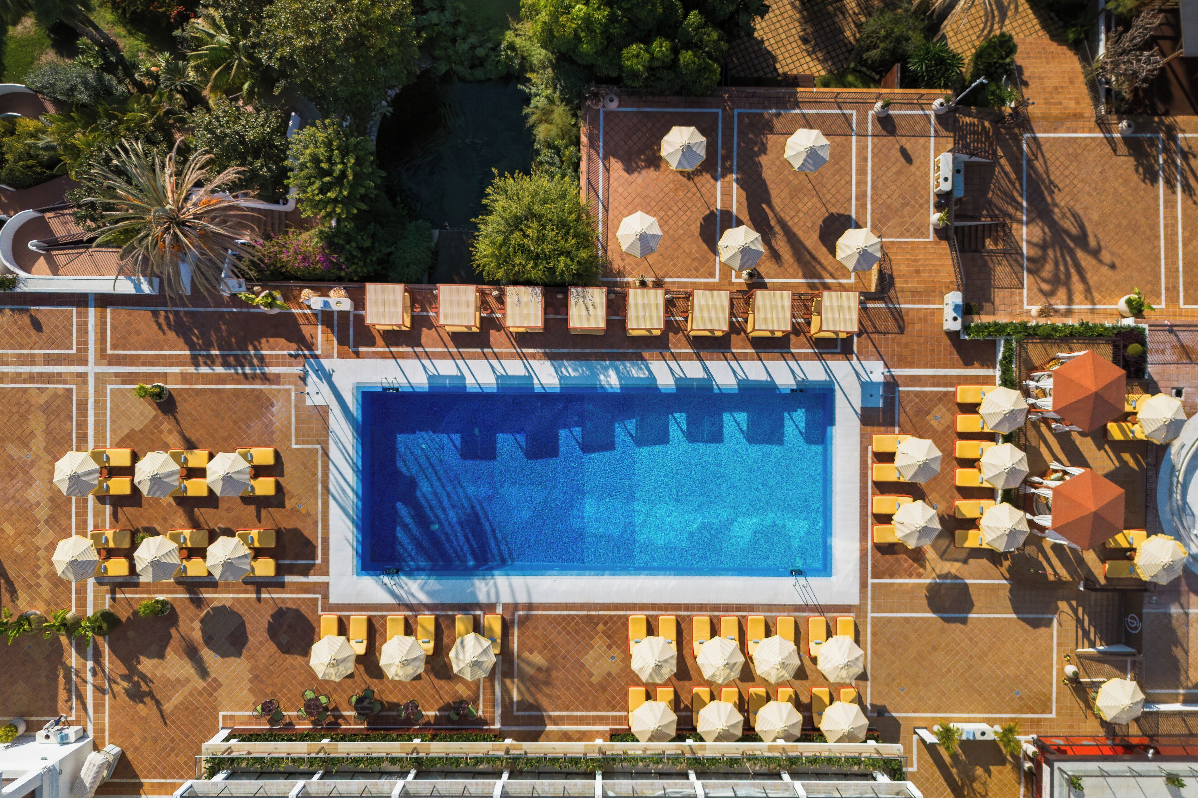 Vista aérea de un hotel con una gran piscina rodeada de tumbonas y sombrillas, y follaje y árboles al fondo.