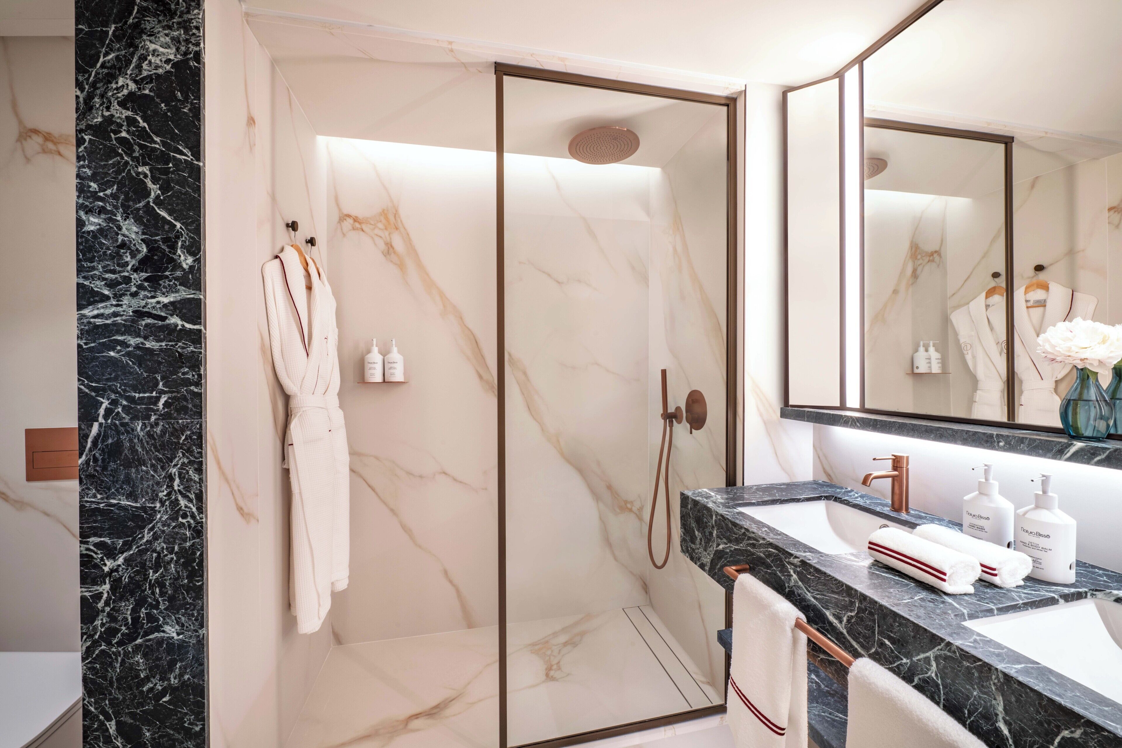 Un baño moderno con pared de mármol, ducha a ras de suelo, espejo grande y encimera de mármol con artículos de aseo.