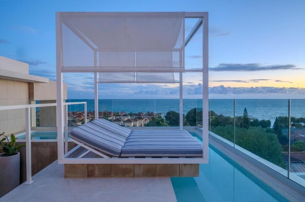 A balcony with a white canopy and a cushioned bench overlooking a pool and a coastal view.