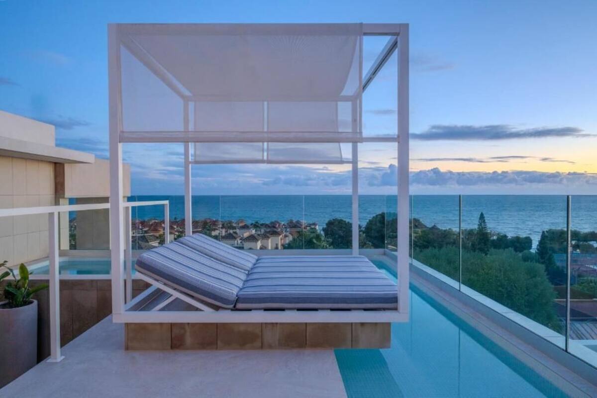 A balcony with a white canopy and a cushioned bench overlooking a pool and a coastal view.