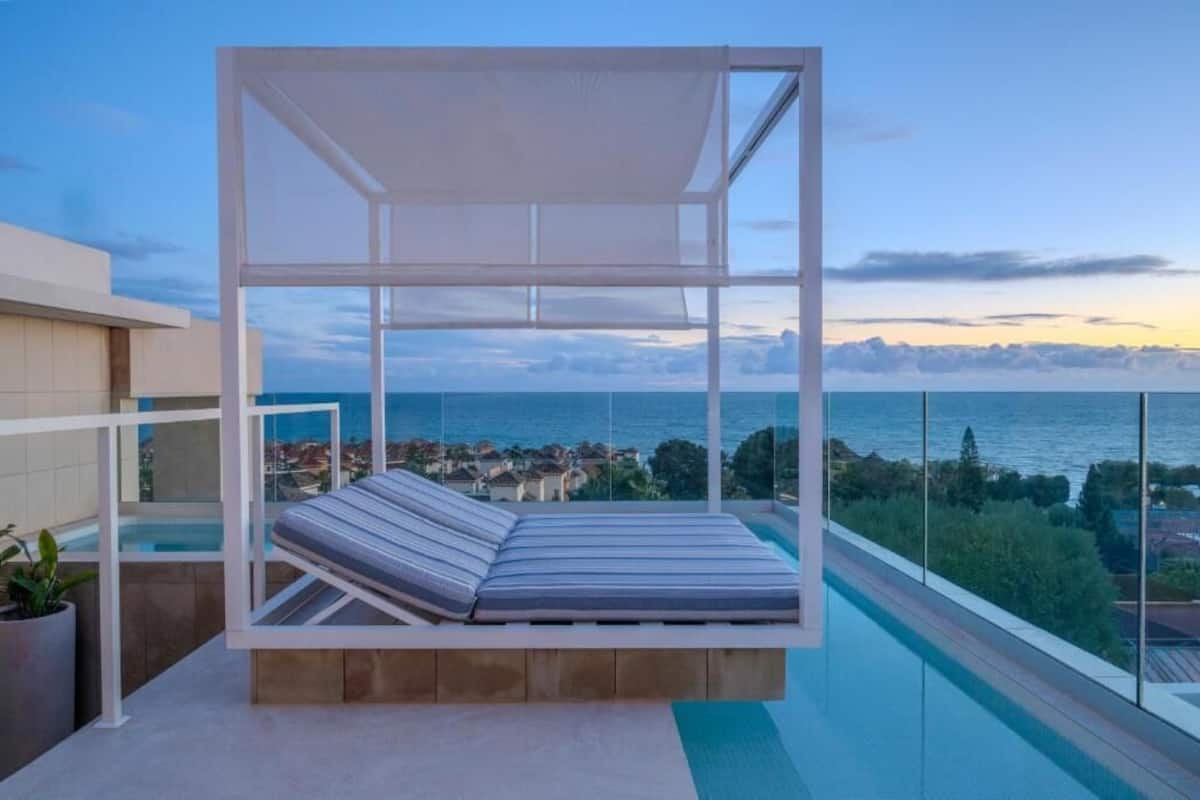 A balcony with a white canopy and a cushioned bench overlooking a pool and a coastal view.