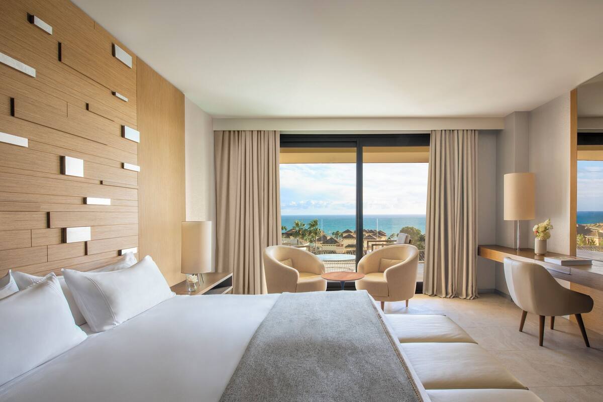 A hotel room with a bed, two chairs, a desk, and a large window with a view of the ocean.