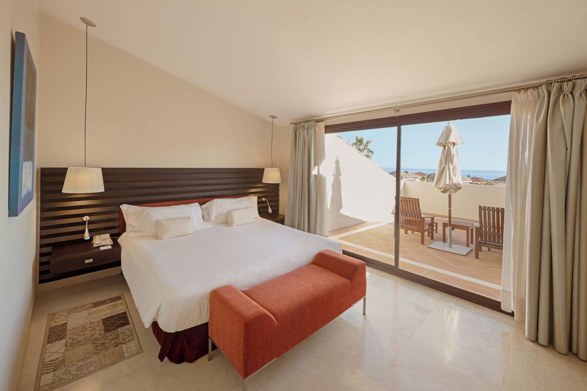 A hotel room with a large bed, a bedside table with a lamp, a small rug, and a balcony with outdoor furniture.