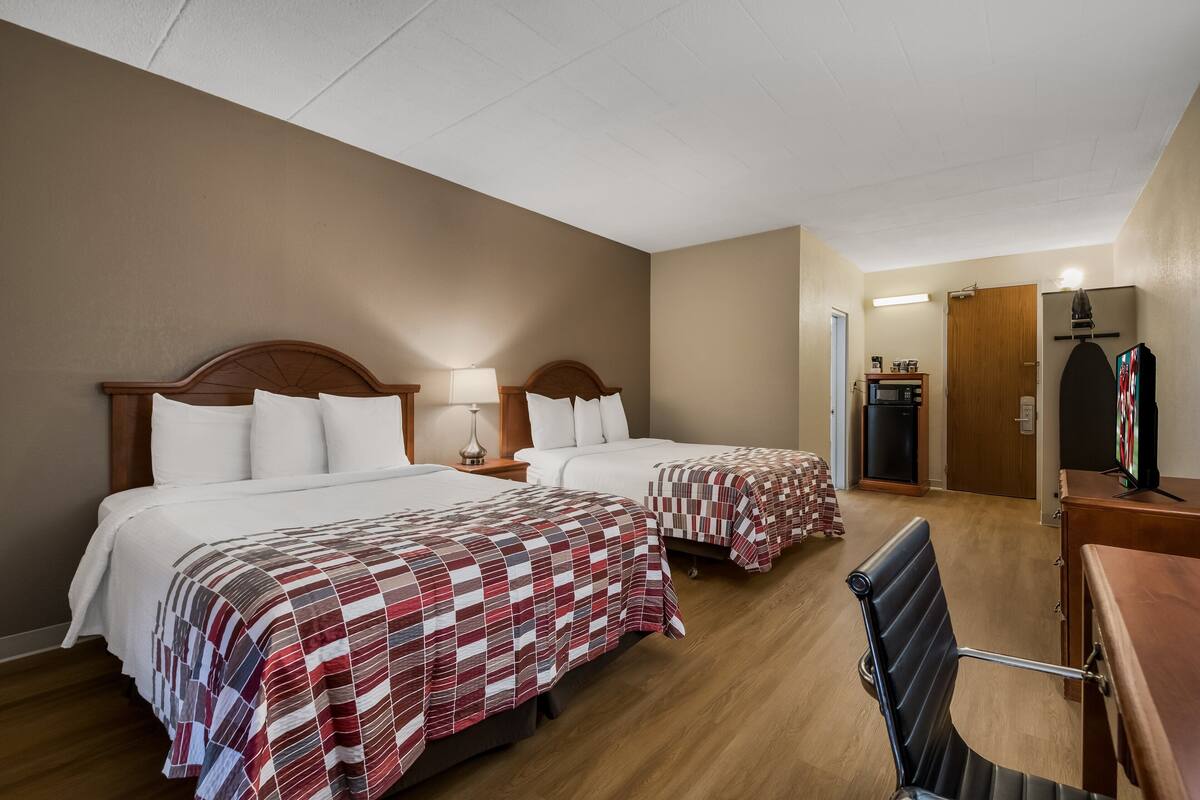 A hotel room with two beds, a desk, and a chair. There is a wooden dresser and a door leading to another room.