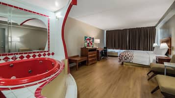 A hotel room with a red jacuzzi, a bed with a patterned bedspread, a desk with a lamp, and a large mirror.