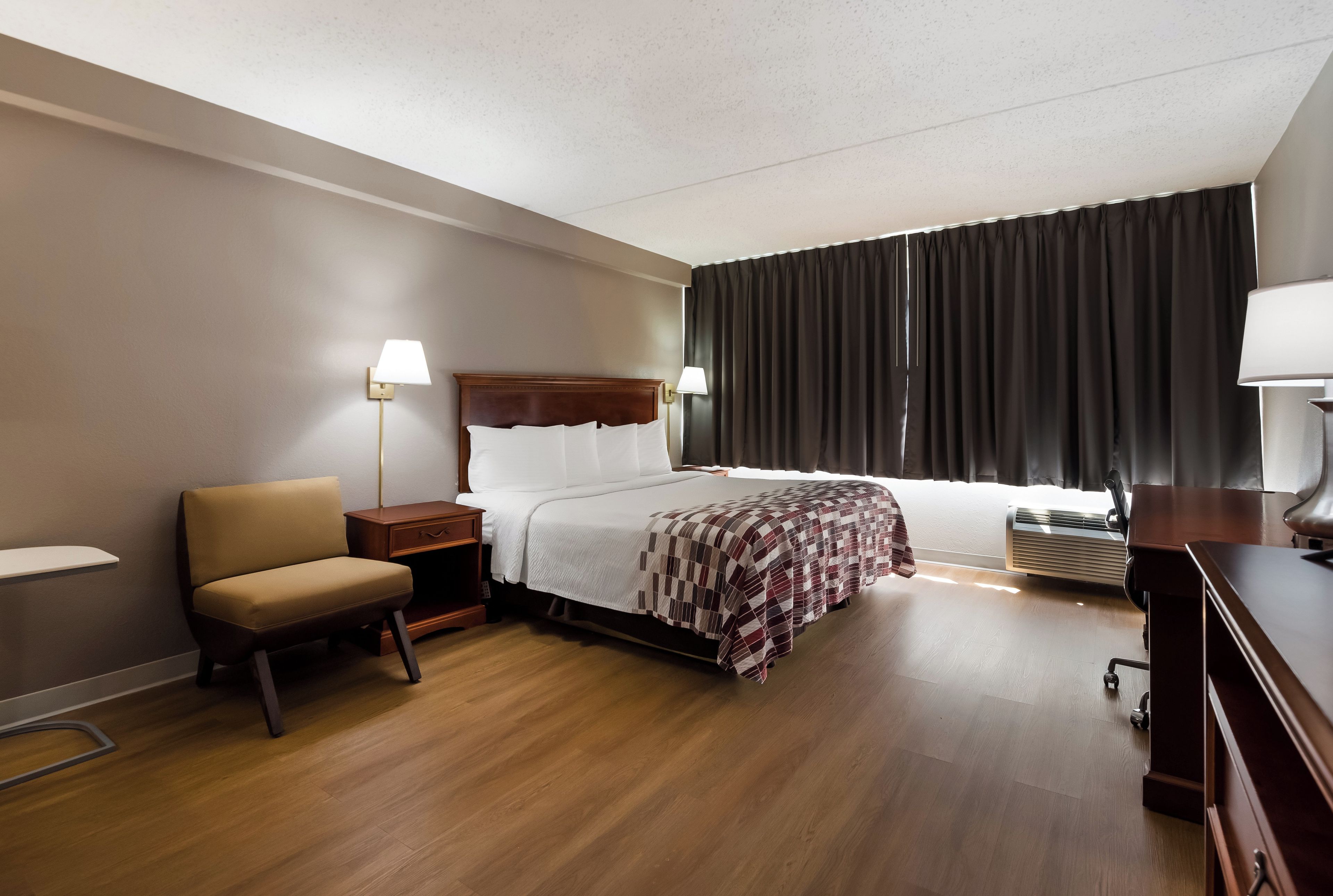A hotel room with a bed, a chair, a bedside table, a lamp, a window with curtains, and a radiator.