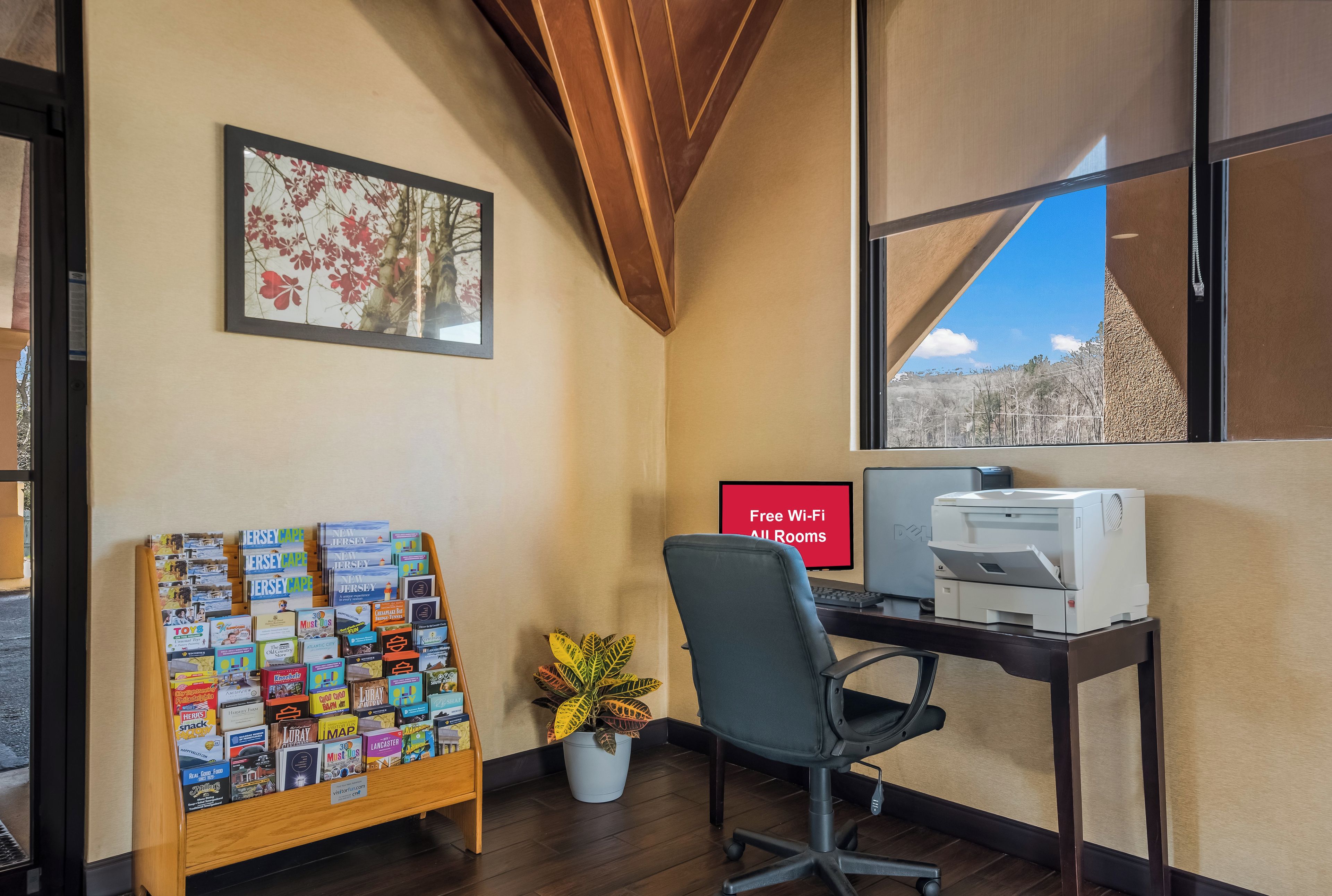 A hotel room with a desk, a computer, a printer, a chair, a plant, a bulletin board with brochures, a framed picture, and a window with a view.
