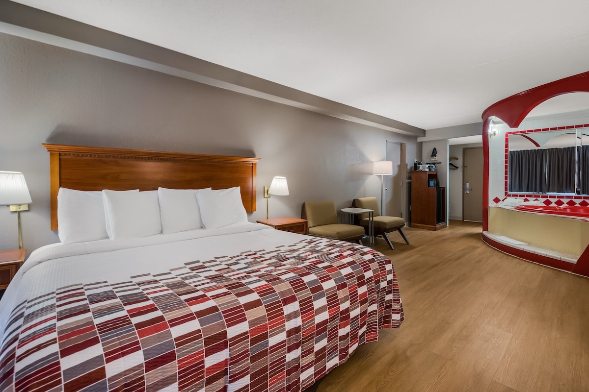 Habitación de hotel con una cama grande, un escritorio, dos sillas, un espejo y un baño visible a través de un arco.