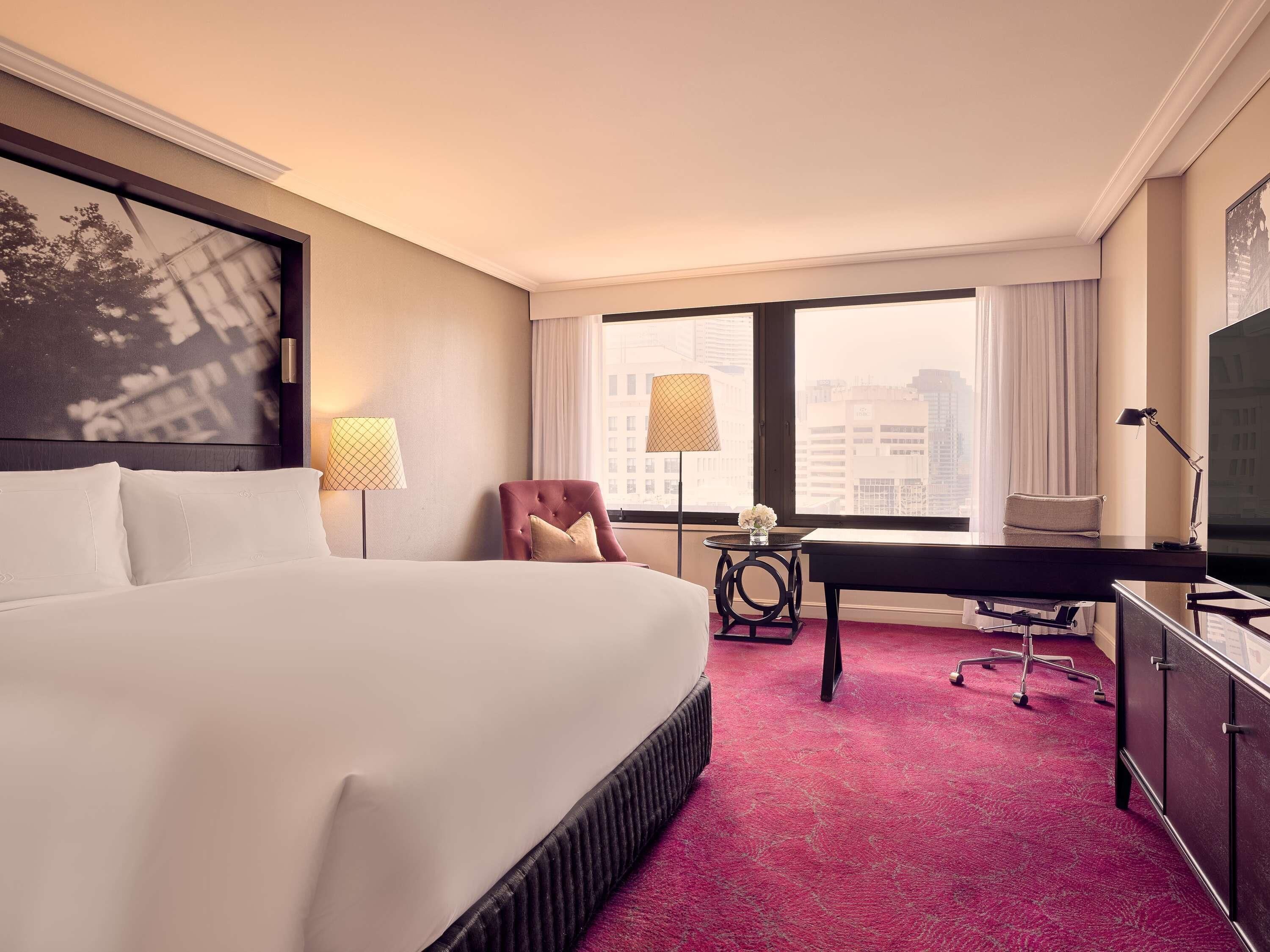 A hotel room with a large bed, a desk, a chair, a TV, and a city view through the window.