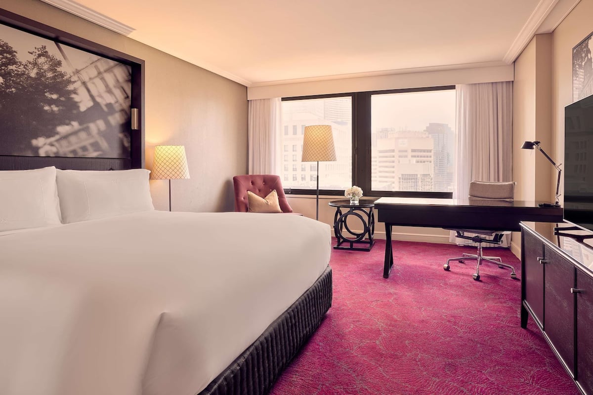 A hotel room with a large bed, a desk, a chair, a TV, and a city view through the window.