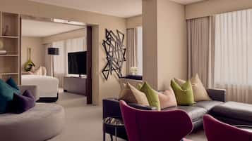 A modern hotel room with a sofa, armchairs, a TV, and a decorative wall piece.
