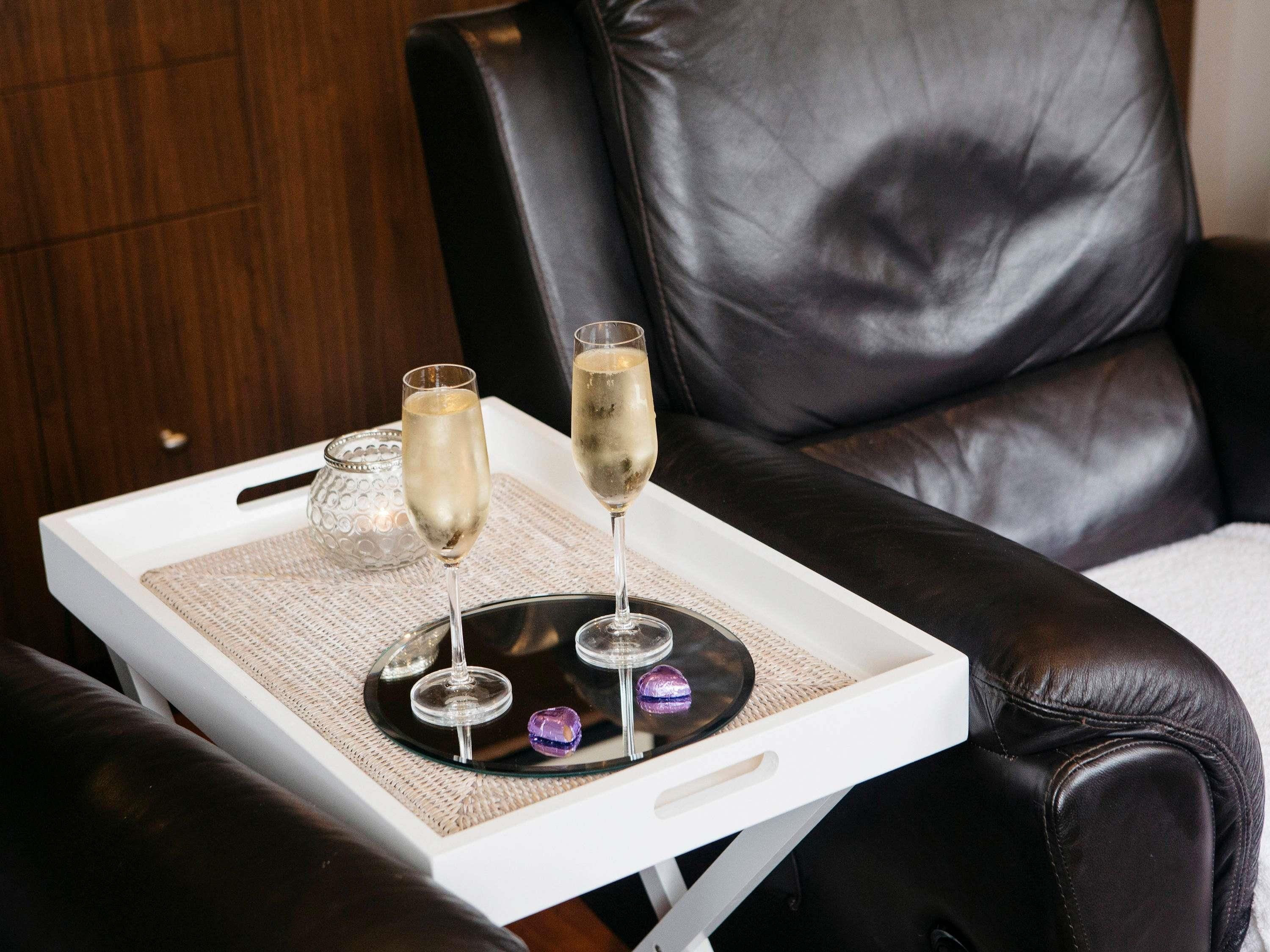 A tray with two champagne glasses and a glass container on a white tray, placed on a chair with a woven placemat.