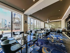 A hotel room with large windows, a city view, a dining area with tables and chairs, and a carpeted floor. - Sofitel Brisbane Central (Brisbane)