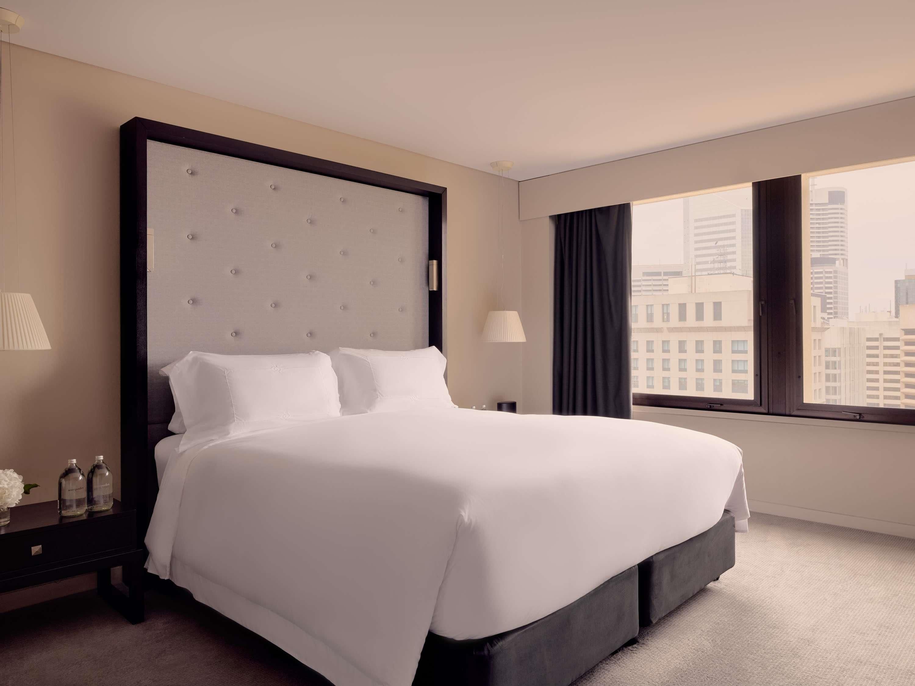 A hotel room with a large bed, a nightstand, a lamp, a window with a city view, and a view of the cityscape.