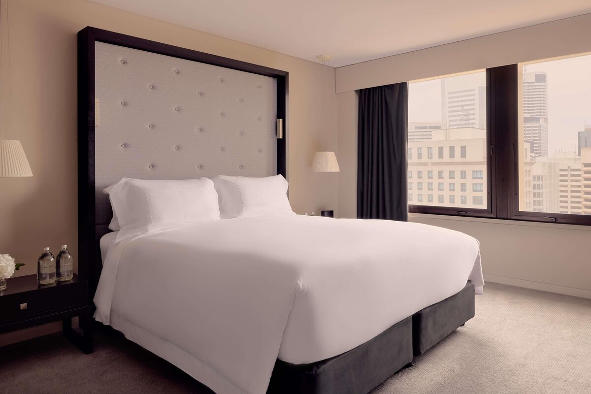 A hotel room with a large bed, a nightstand, a lamp, a window with a city view, and a view of the cityscape.