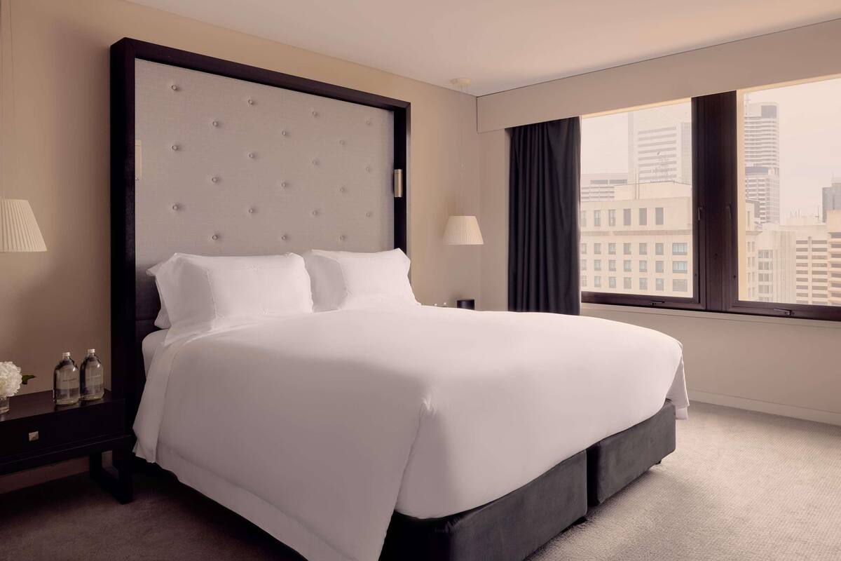 A hotel room with a large bed, a nightstand, a lamp, a window with a city view, and a view of the cityscape.
