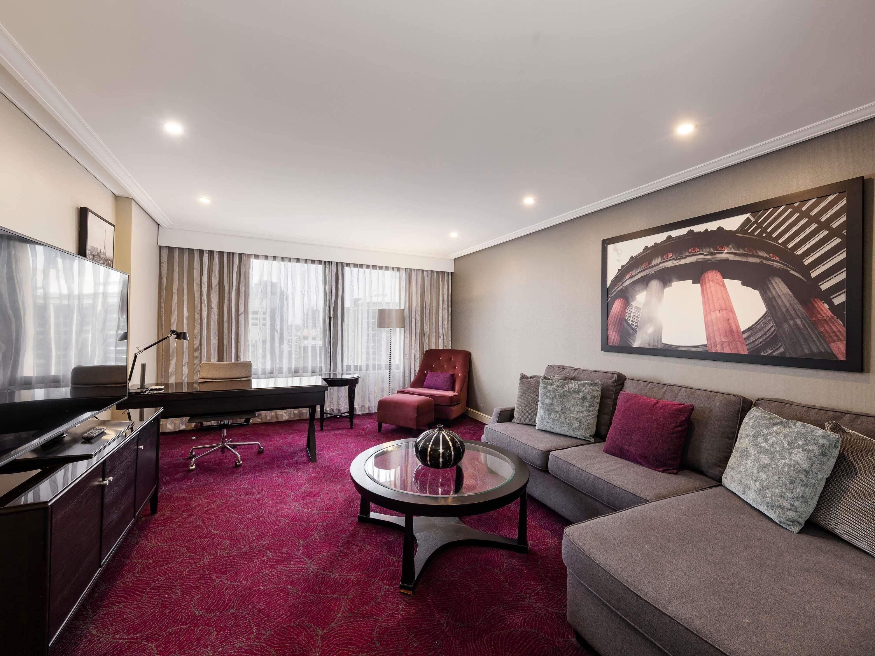 A hotel room with a sofa, armchair, TV, desk, and a painting of a bridge.