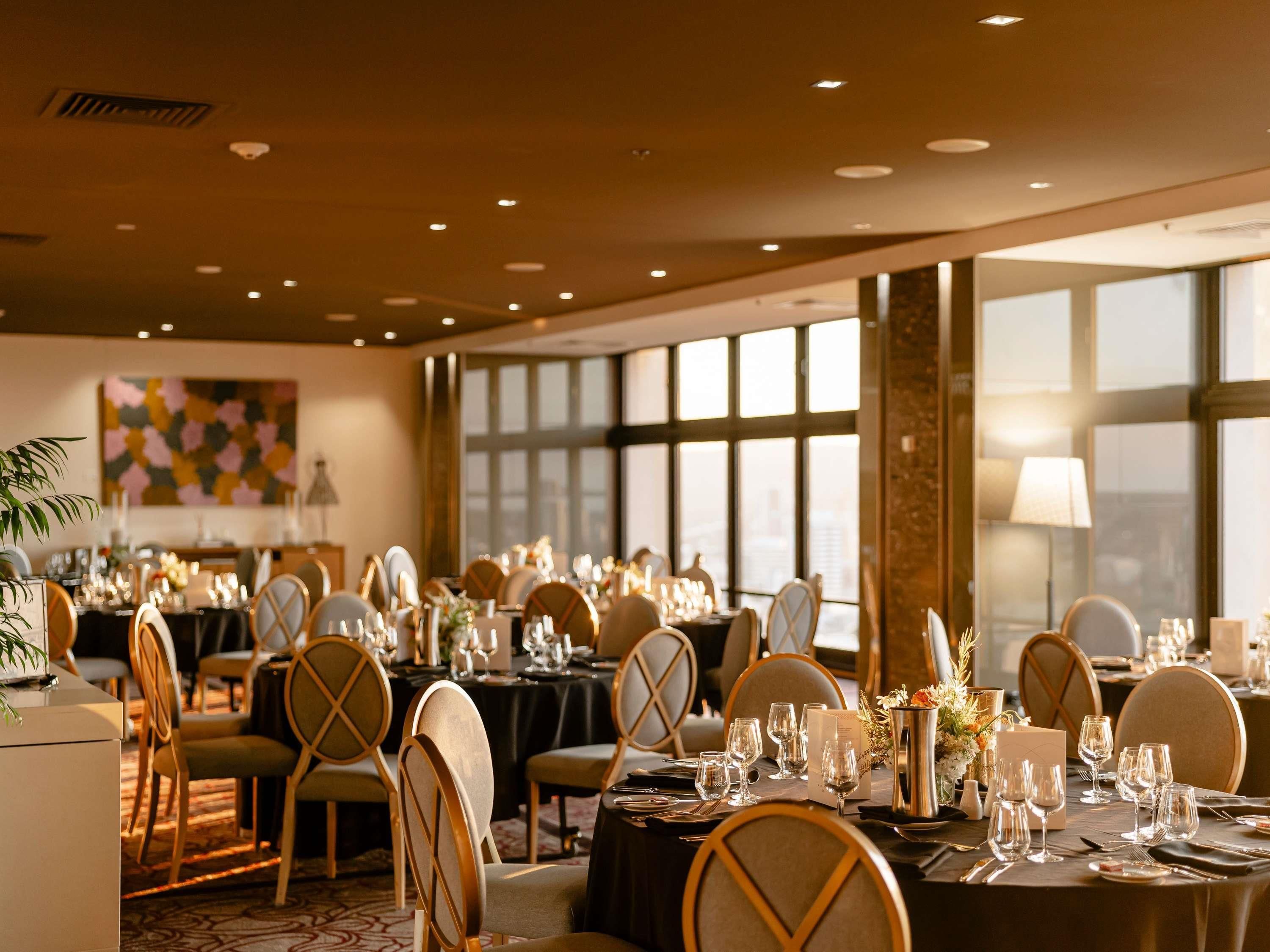 A well-lit banquet hall with round tables, chairs, and a large window providing a view of the city.
