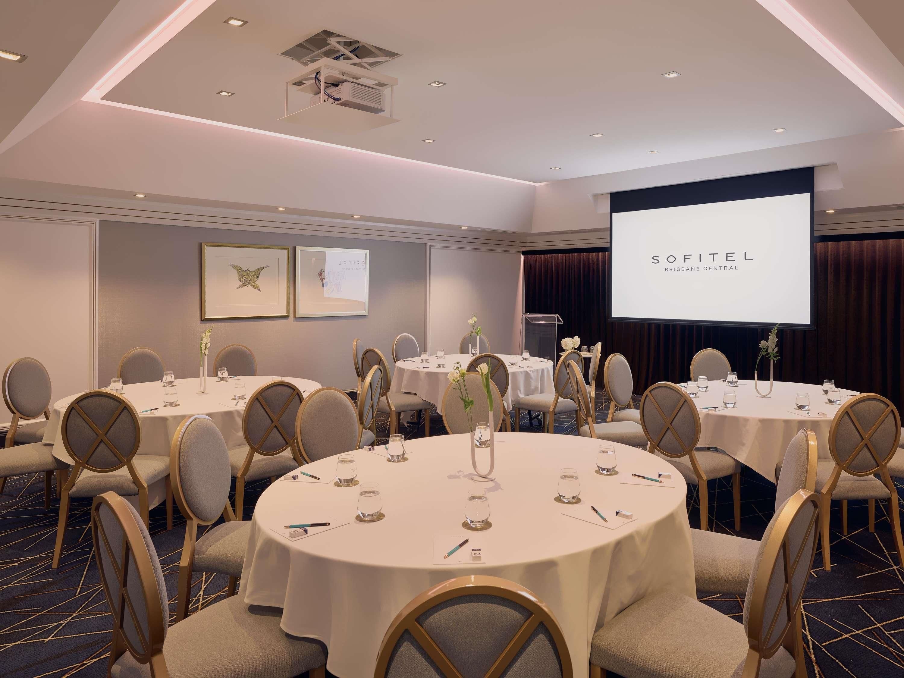A conference room with round tables, chairs, and a projection screen displaying 'SOFITEL BRISBANE CENTRAL'.