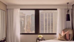 Habitación de hotel con un ventanal grande, una cama, una mesita con un jarrón de flores y un sillón con cojines.