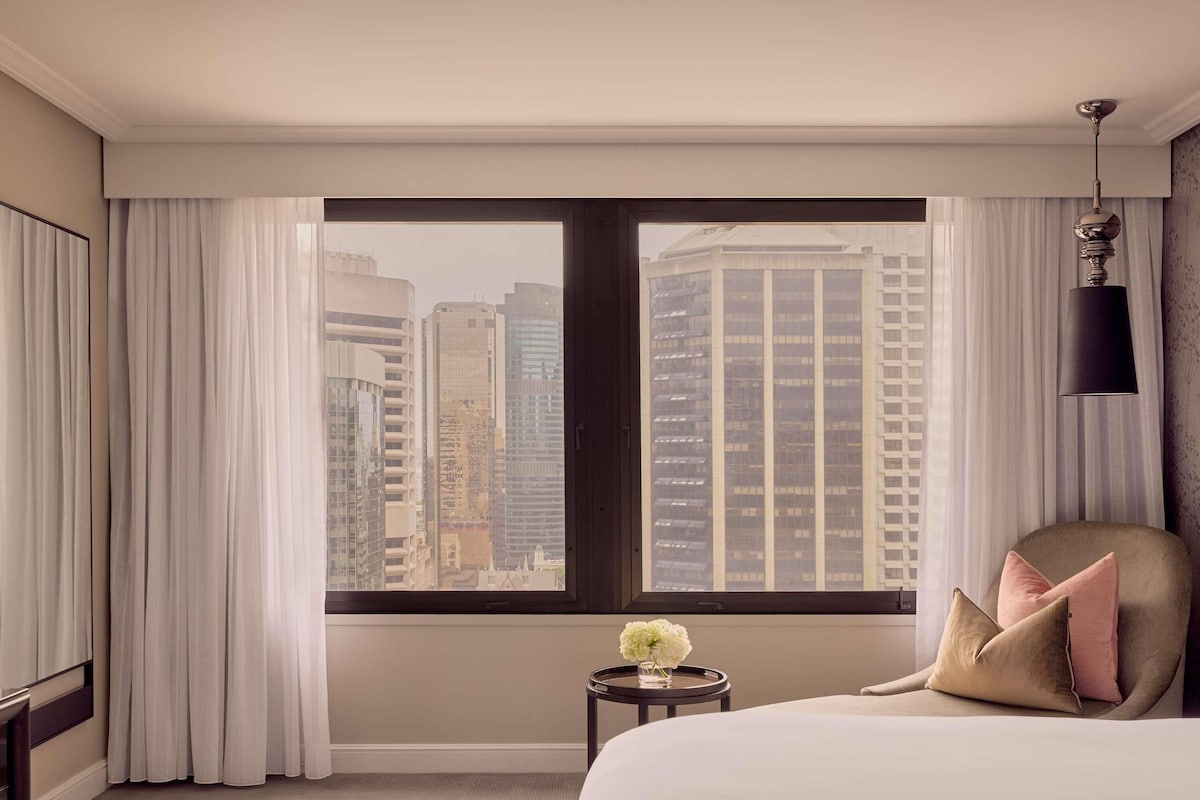 A hotel room with a large window, a bed, a small table with a vase of flowers, and a chair with cushions.